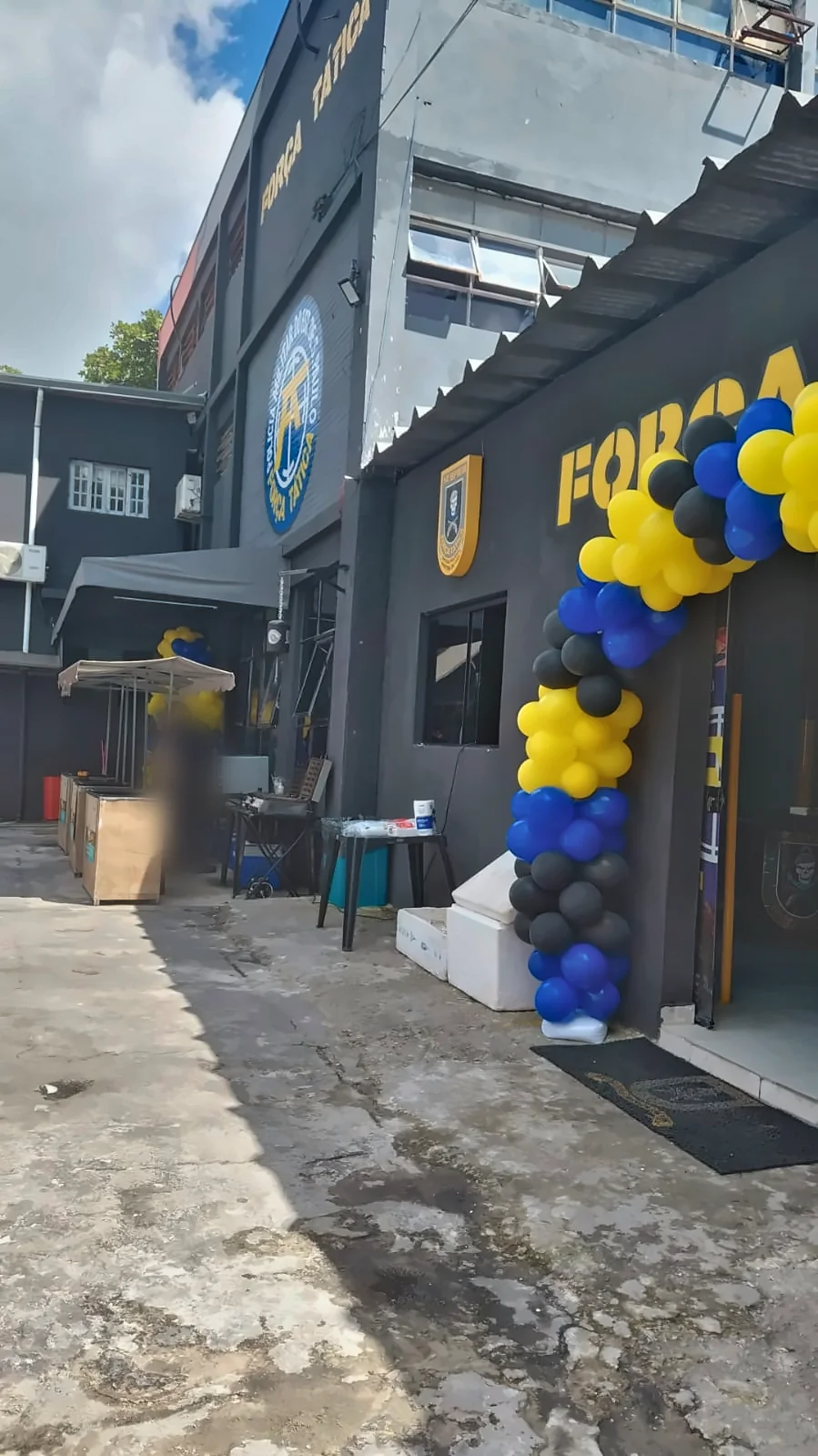 Entrada da unidade da Força Tática decorada com arco de balões azuis, amarelos e pretos e as barraquinhas montadas ao fundo